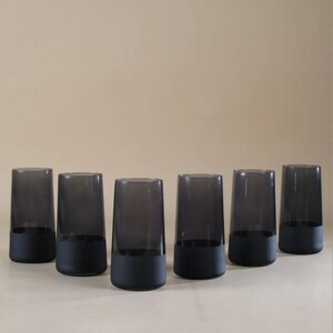 May include: Six black glass tumblers with a frosted bottom. The tumblers are arranged in a semi-circle on a light beige surface.