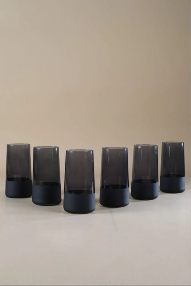May include: Six black glass tumblers with a frosted bottom. The tumblers are arranged in a semi-circle on a light beige surface.