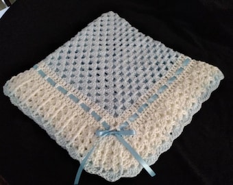 May include: A handmade, crocheted baby blanket in soft blue and cream colors. The blanket features a delicate openwork design with a scalloped edge and is adorned with light blue satin ribbon accents and a small bow.