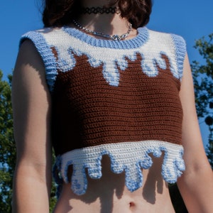 Puede incluir: Un top corto de crochet marrón, blanco y azul con un diseño de goteo. El top es sin mangas y tiene un escote redondo.