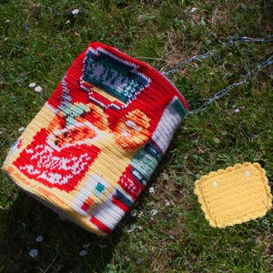 Könnte beinhalten: Eine farbenfrohe, gehäkelte Tasche mit Kettenriemen liegt auf grünem Gras neben einem kleinen, gelben gehäkelten Quadrat. Die Tasche hat rote, gelbe, weiße und grüne Muster. Die Kette ist silberfarben.
