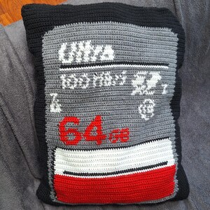 Peut inclure: Un coussin crocheté en forme de carte mémoire. Il présente un fond gris avec des accents blancs et rouges, et le texte "ULTRA 100 MB/s" et "64 GB". La bordure est noire.