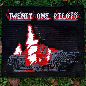Peut inclure: Couverture noire au crochet avec l'inscription "TWENTY ONE PILOTS" en blanc et rouge. En dessous, un motif stylisé en rouge, blanc et gris. La couverture est posée sur de l'herbe verte avec des feuilles mortes.