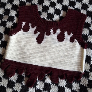 May include: A white crocheted sleeveless top with a maroon crocheted neckline and bottom trim. The maroon trim has a dripping effect.