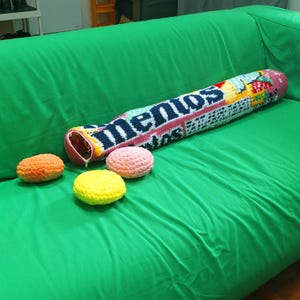 May include: A green sofa with a large, crocheted Mentos candy roll pillow and three crocheted candies in orange, pink, and yellow. The pillow has the word "Mentos" in navy blue.