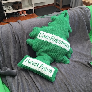 May include: A green, crocheted car air freshener in the shape of a Christmas tree, with the words "Car-FRESHNER" and "Forest Fresh" in white. The freshener is lying on a gray blanket.