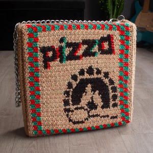 May include: A handmade, square crochet bag with a chain strap. The bag features the word "pizza" in black and red, along with a black graphic. The edges are trimmed with red and green stitching. The bag is tan.