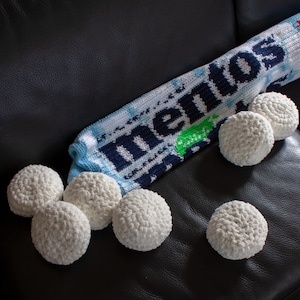 May include: A crocheted Mentos candy roll and several white crocheted balls are arranged on a dark surface. The Mentos roll is blue, white, and green, with the word "Mentos" in dark blue. The balls are round and textured.