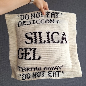 Peut inclure: Coussin en crochet de couleur crème avec les mots "SILICA GEL" en grandes lettres noires. Le texte "DO NOT EAT" et "THROW AWAY" est également affiché. Le coussin est tenu par une personne.