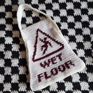 Peut inclure: Sac en crochet blanc avec un panneau d'avertissement rouge indiquant "WET FLOOR". Le sac a une poignée et est posé sur une surface à carreaux noirs et blancs.