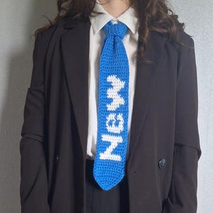 Peut inclure: Une cravate en crochet bleu avec le mot "Meow" écrit en blanc. La cravate est portée par une personne portant une chemise blanche et un blazer noir.
