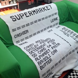 Peut inclure: Une couverture en crochet blanc avec un motif graphique noir représentant un ticket de caisse de supermarché. Le texte inclut SUPERMARKET, CASHIER, NAME, et une liste d'articles comme CANDY ROLL, SMILEY POTATO, DETERGENT PODS, SD CARD, ALPHABET PASTA, RAVIOLI CAN et BUTTER.