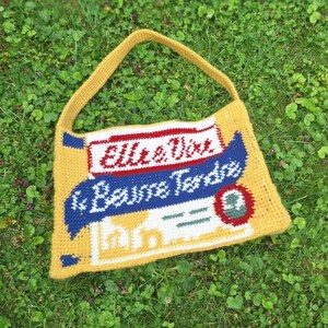 Peut inclure: Sac fourre-tout crocheté à la main, de couleur jaune, avec un motif comportant le texte "Elle & Vire & Beurre Tendre" en rouge, blanc et bleu. Le sac est doté d'une longue bandoulière jaune et posé sur de l'herbe verte.