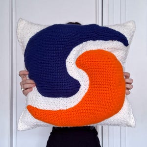 May include: A handmade crochet pillow with a white background and a large, abstract design in navy blue and orange. The pillow is square-shaped and appears to be soft and plush, perfect for home decor.