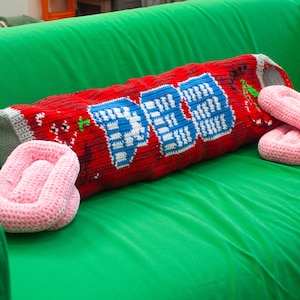 Peut inclure: Un coussin cylindrique en crochet rouge arborant le texte PEZ en blanc et bleu, avec des extrémités grises et de petits motifs de cerises rouges. Deux objets ovales en crochet rose clair sont placés de chaque côté du coussin cylindrique.