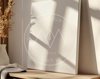 May include: A white canvas mockup with a white circle around it that says "MOCKUP STUDIO DELIGHT LAND" with a pen and mountain design inside the circle. The canvas is sitting on a wooden surface with a white vase and a beige throw blanket.