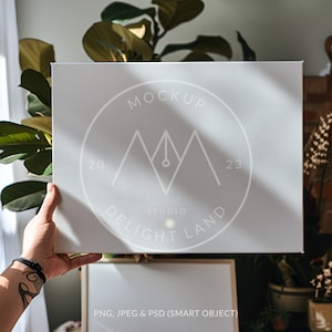 May include: A light gray canvas mockup held by a person's hand. The canvas features a circular logo with the text "MOCKUP STUDIO DELIGHT LAND 2023". The bottom of the image includes the text "PNG, JPEG & PSD (SMART OBJECT)".