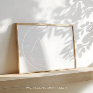 Pode incluir: Uma maquete de moldura branca com uma moldura de madeira clara, sentada em uma prateleira de madeira clara. A moldura está em frente a uma parede branca com sombras de uma planta.