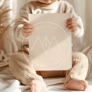 May include: A baby in a beige sweater and pants holds a blank white card with the text "MOCKUP" and "STUDIO DELIGHT LAND" printed on it.