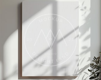 Könnte beinhalten: Ein weißer Leinwand-Mockup mit einem weißen Kreislogo, auf dem "MOCKUP STUDIO DELIGHT LAND" steht, mit einem stilisierten Bergdesign und einem Stift im Kreis. Die Leinwand hängt an einer weißen Wand mit einem Schatten von einem Fenster.