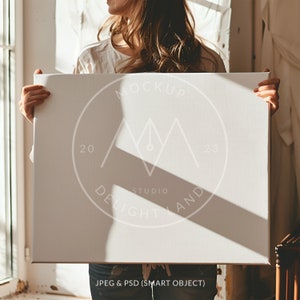 Puede incluir: Una maqueta de lienzo blanco con un logotipo circular blanco que dice "MOCKUP STUDIO DELIGHTLAND 20 23" en una fuente sans-serif. El lienzo está siendo sostenido por una persona frente a una ventana.