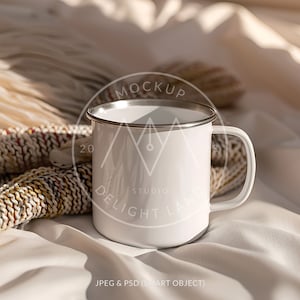 Könnte beinhalten: Weiße Emaille-Campingtasse mit silbernem Rand und Henkel. Die Tasse steht auf einer weißen und beigefarbenen Decke mit einer gestrickten Wurfdecke.