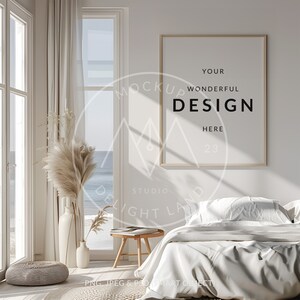 May include: A white bedroom with a large window overlooking the ocean. The bed is made with white linens and a throw pillow. A wooden side table with a book and a vase of dried flowers sits next to the bed. A framed poster with the text "YOUR WONDERFUL DESIGN HERE 23" hangs on the wall.