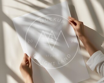 Puede incluir: Una hoja de papel blanca con el texto "MOCKUP" y "STUDIO DELIGHT LAND" en un círculo. El papel está siendo sostenido por las manos de una persona. El fondo es una superficie beige claro con sombras de una ventana.