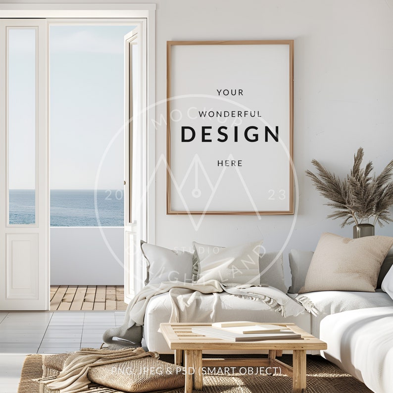 May include: A white couch with beige pillows in a living room with a large window overlooking the ocean. A wooden coffee table with books on it sits in front of the couch. A framed poster with the text "YOUR WONDERFUL DESIGN HERE" hangs on the wall.