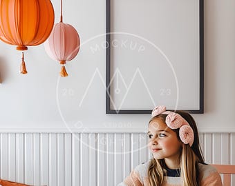 Può includere: Una bambina con un maglione a righe rosa e bianco e una fascia per capelli in crochet rosa è seduta a un tavolo con uno sfondo bianco. Una cornice nera con uno sfondo bianco è appesa al muro dietro di lei. Due lanterne di carta pendono dal soffitto sopra il tavolo.