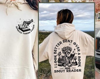 May include: A beige hoodie with a black graphic print on the front that says "Spread those pages" and a black graphic print on the back that says "I look better bent over a book, Proud Smut Reader".