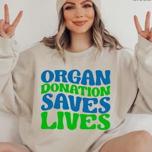 May include: A light beige sweatshirt with the text "ORGAN DONATION SAVES LIVES" in blue and green letters. The letters are in a retro, rounded font.