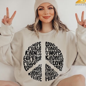 May include: A beige sweatshirt with a peace sign design. The peace sign is made up of the words "Hope", "Peace", "Love", and "Kindness" repeated in a circular pattern.