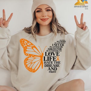 May include: A beige sweatshirt with an orange butterfly graphic and the text "MULTIPLE SCLEROSIS IS A JOURNEY...LOVE LIFE HATE THE DISEASE AND FIGHT." The sweatshirt also features a small orange ribbon.