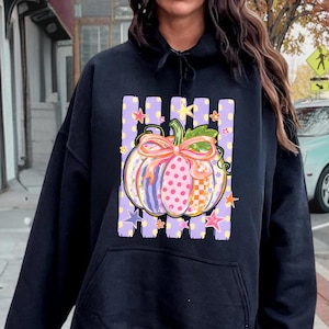 May include: A black hooded sweatshirt featuring a graphic print of a pumpkin with a pink bow. The pumpkin has a multi-colored pattern of stripes, polka dots, and checkers. The background of the graphic consists of vertical purple stripes and stars.