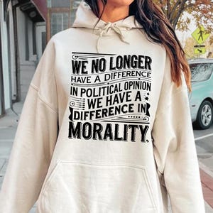 May include: A sand-colored hoodie with a front pocket and drawstring hood. The front of the hoodie features the text: "WE NO LONGER HAVE A DIFFERENCE IN POLITICAL OPINION WE HAVE A DIFFERENCE IN MORALITY."
