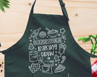 May include: A dark green apron with the words "Christmas Baking Crew" and baking-themed illustrations in white. The apron has two pockets and a neck strap. The background includes cooking utensils and ingredients.