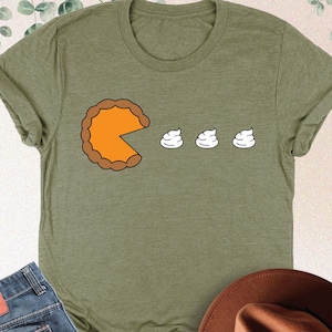 May include: A green t-shirt with a graphic of a pumpkin pie with a bite taken out of it and three dollops of whipped cream. The text "HEATHER OLIVE" is at the top of the image.