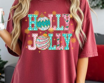 May include: Burgundy t-shirt with the words "HOLLY JOLLY" in a colorful, festive font, with ornament graphics. A white mug with a Christmas tree design is held in the left hand. A Christmas tree and wrapped gifts are in the background.