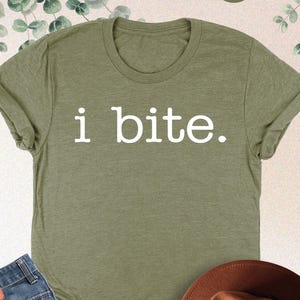 May include: Heather olive green t-shirt with the text "i bite." paired with blue jeans and a brown fedora hat. The shirt is a casual style, suitable for everyday wear.