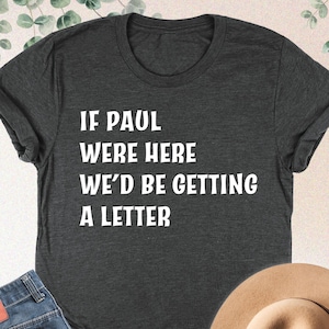 May include: Dark grey heather t-shirt with the text "IF PAUL WERE HERE WE'D BE GETTING A LETTER" in white. The shirt is paired with blue jeans and a tan hat. The image also includes a sprig of green leaves.