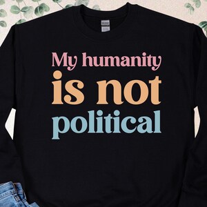 May include: Black crewneck sweatshirt with the text "My humanity is not political" in pink, orange, and blue. The sweatshirt is paired with blue jeans and black and white sneakers.