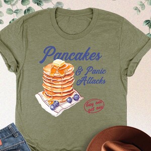 May include: A green t-shirt with a graphic of a stack of pancakes with syrup and blueberries. The text "Pancakes & Panic Attacks" is printed on the shirt. The text "buy one get one" is also printed on the shirt.