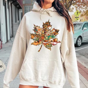 May include: A sand-colored hoodie featuring a large, colorful maple leaf design. The leaf is composed of various autumn-themed patterns, including pumpkins, plaid, and floral prints. The hoodie has a front pocket and a drawstring hood.