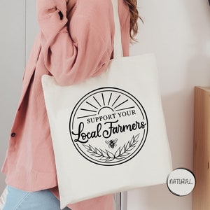 May include: A white canvas tote bag with black text that reads "Support Your Local Farmers" in a circle with a sun, wheat, and a bee. The bag is being held by a person wearing a pink sweater and blue jeans.