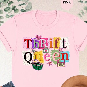 May include: Pink t-shirt with the words "Thrift Queen" in a colorful design. The image also includes a pair of blue jeans and a brown hat. The words "Second Hand" are also visible.