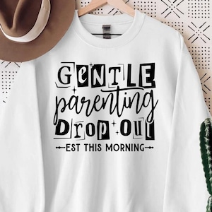 May include: A white crewneck sweatshirt featuring black text that reads GENTLE PARENTING DROP OUT EST THIS MORNING, accompanied by small star accents. The sweatshirt is displayed on a wooden hanger alongside a brown fedora hat.