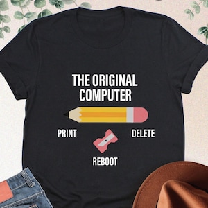 May include: A black t-shirt with a graphic of a pencil and the text "The Original Computer". The pencil has a pink eraser and the text "Print", "Reboot", and "Delete" are below the pencil.