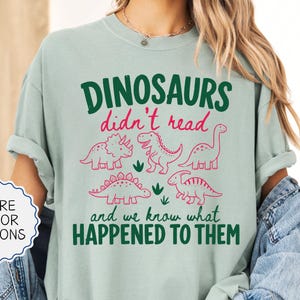 May include: A light green t-shirt featuring a graphic with the text "DINOSAURS didn't read and we know what HAPPENED TO THEM" in dark green and pink. The design includes line art illustrations of five different dinosaurs in pink ink.
