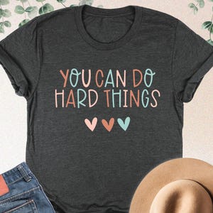 May include: Dark grey heather t-shirt with the text "YOU CAN DO HARD THINGS" in colorful letters and three hearts. The image also includes blue jeans and a tan hat.